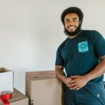 A cheerful man with afro hair standing next to moving boxes indoors, smiling.