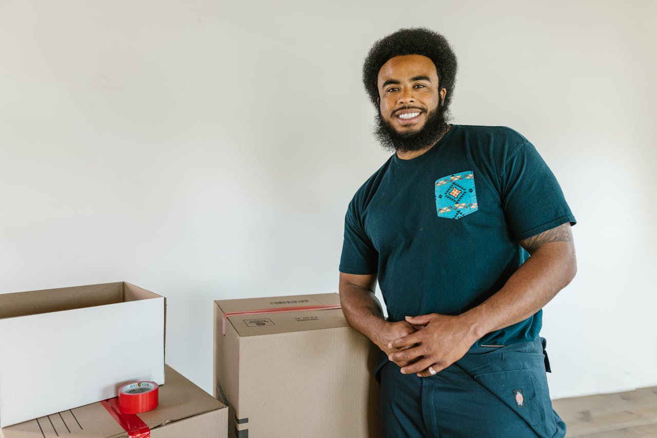 home-img A cheerful man with afro hair standing next to moving boxes indoors, smiling.